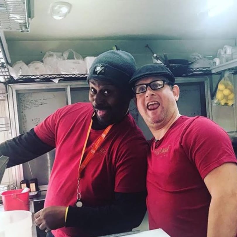 Two male Succotash kitchen staff in red shirts and hats smiling playfully at the camera.