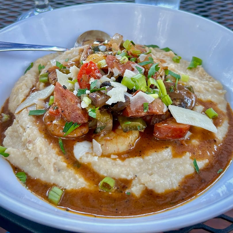 Succotash's shrimp and grits, topped with sausage, peppers, and green onions, in a white bowl.