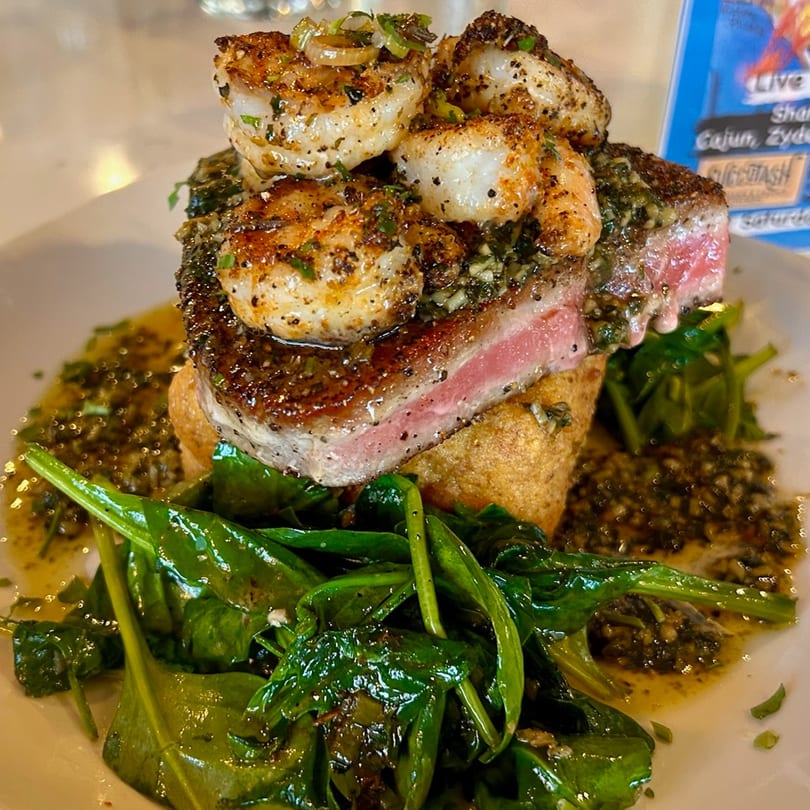 Seared tuna steak topped with seasoned shrimp, served over greens and a savory sauce.