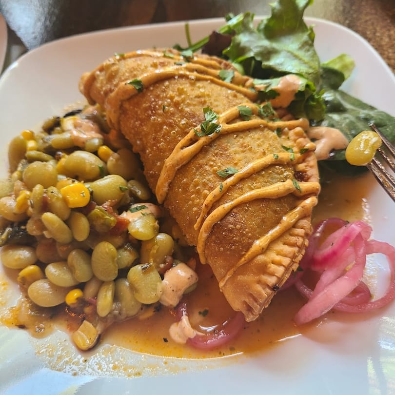 A golden-brown empanada drizzled with sauce, served with lima beans, corn, and pickled red onions.