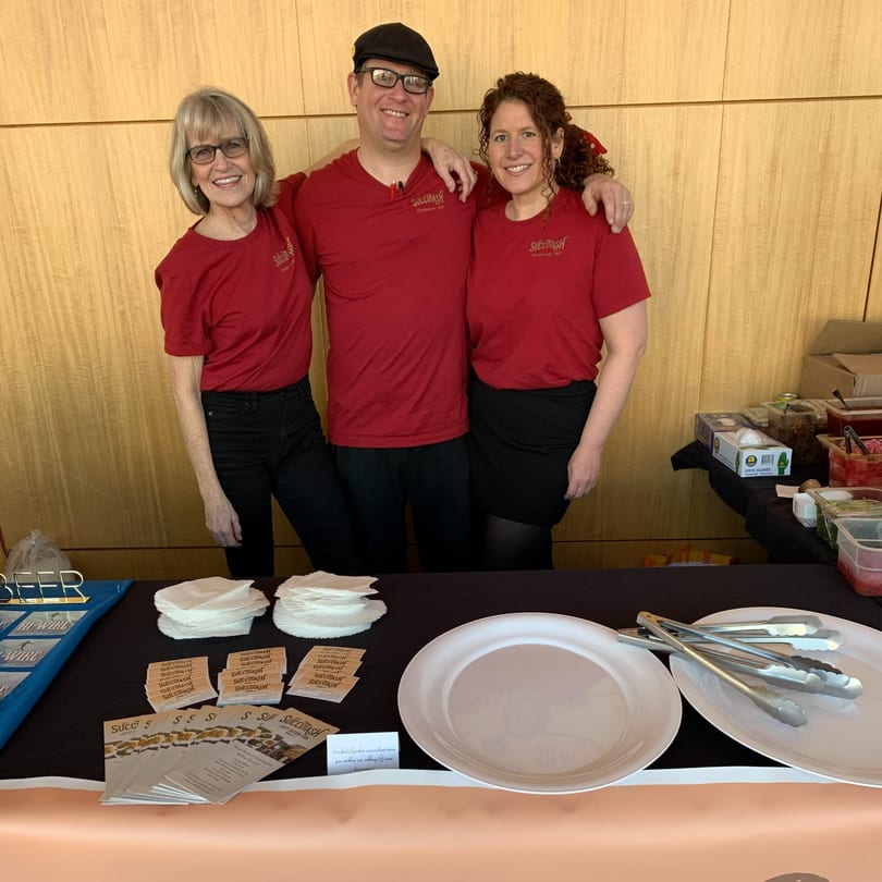 Three smiling Succotash staff in red shirts stand behind a table with plates and brochures.