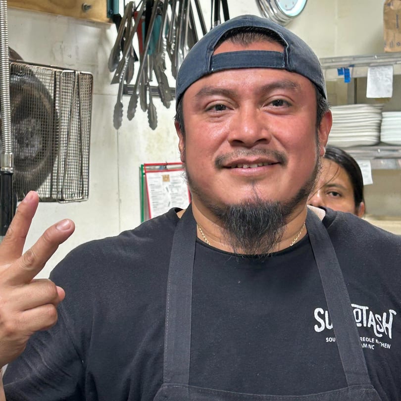A smiling Succotash chef in a black apron and cap gives a peace sign in the kitchen.