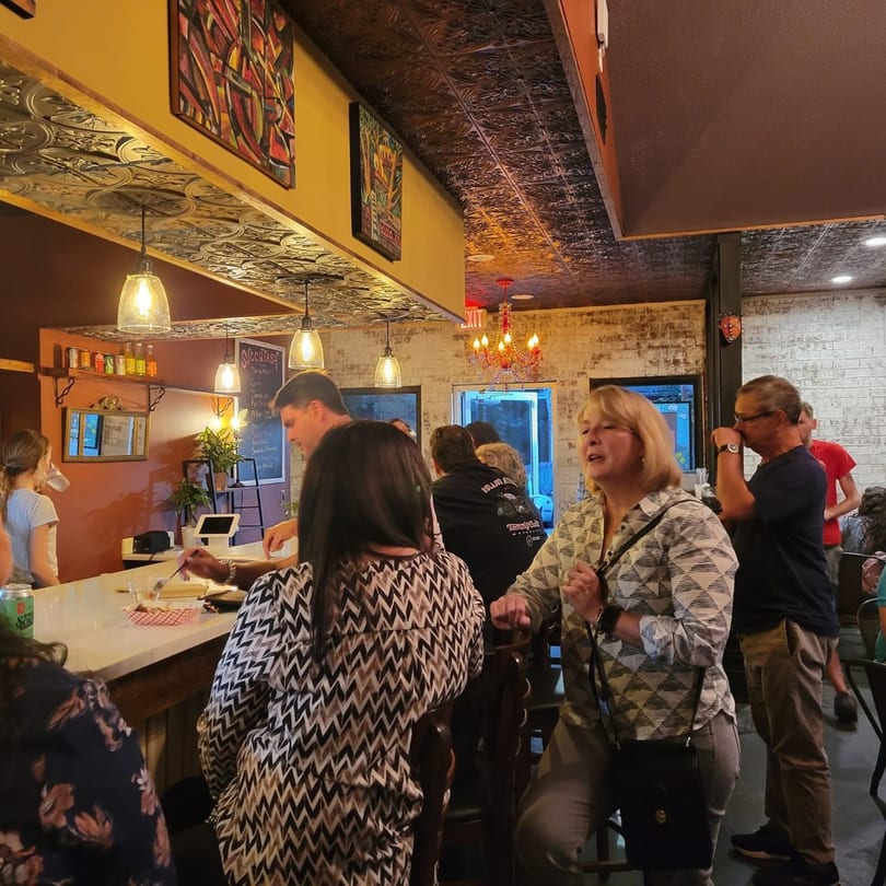 Patrons socialize at the bar of Succotash Southern and Creole Kitchen, featuring a tin ceiling and warm lighting.