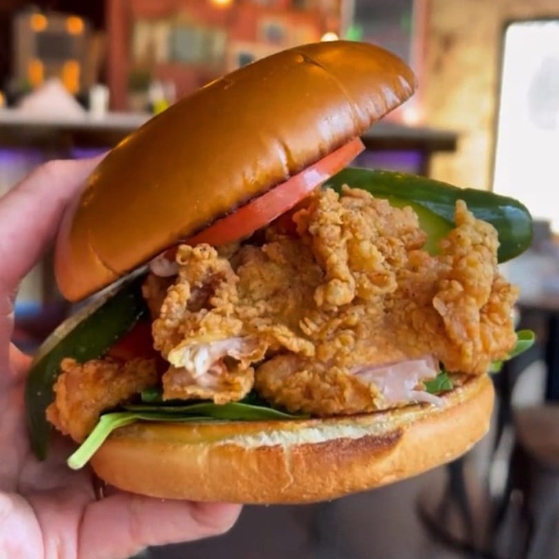 Hand holding a crispy fried chicken sandwich with lettuce, tomato, and pickles on a brioche bun.